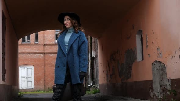 Young Redhead Woman in a Blue Coat and Black Hat with a Suitcase on the Background of the Old City alt