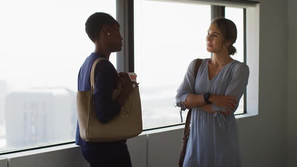 Two diverse female colleagues standing at window, talking and smiling alt