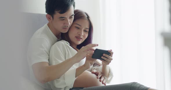 Asian husband and pregnant wife spend time together in the bedroom and using smartphone.