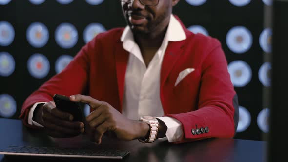 Cute african-american businessman in red suit uses smartphone in dark room.