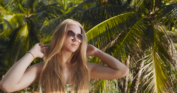 Young Woman with Long Hair and Sunglasses Against Palm Tropical Leaves Posing