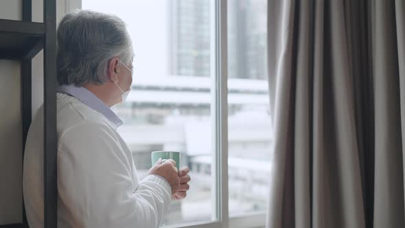 old asian senior retired age standing hand hold hot drink look outside window alt