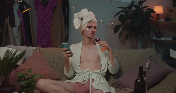 Man Transgender in Loungewear and Undereye Patches Sitting on Couch and Drinking Wine. Young alt