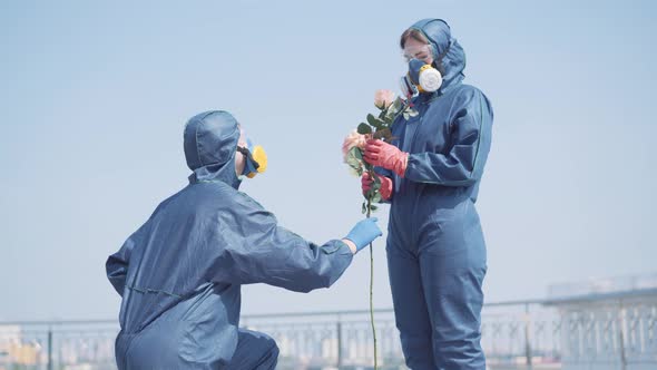 Man Standing on One Knee and Giving Flowers To Woman in Antiviral Suit. Happy Young Caucasian Girl alt