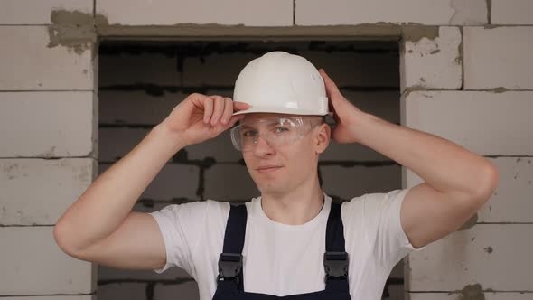 A Male Worker Puts on a White Construction Helmet on a Construction Site alt