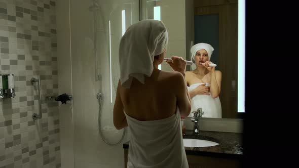 Woman with White Bath Towel on Head Brushing Her Teeth Looking in the Mirror alt