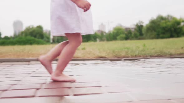 Little Barefoot Girl Running Through Puddles in White Dress in City Park