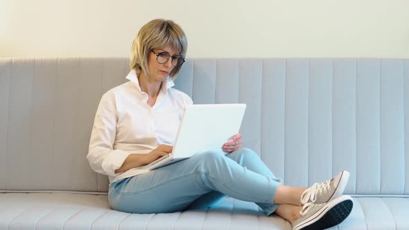 An Elderly Beautiful Woman Works at Home at a Laptop in the Home Office