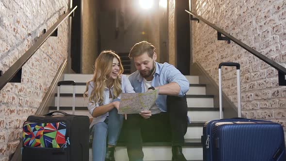 Young Bearded Guy with His Blond Girlfriend which Sitting on the Steps in Hotel and Looking at Map alt