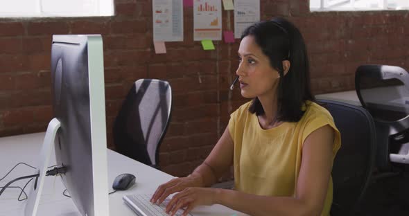 Mixed race businesswoman wearing headset sitting at desk using computer alt