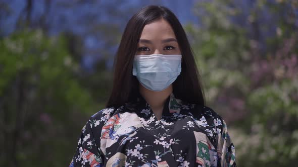 Confident Japanese Woman in Covid Face Mask Looking at Camera Posing in Sakura Garden on Spring Day alt