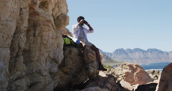 African american man hiking sitting on rock taking photos by the coast alt
