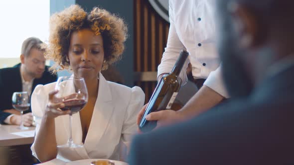 African Woman Taking Red Wine in Modern Restaurant with Waiter Demonstrating Bottle alt
