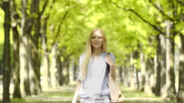 Young Beautiful Blonde Girl in Summer Day Walks at Urban City Park Listening Music with Smartphone