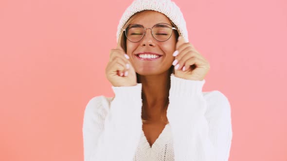 Young Female in Glasses White Jumper and Knitted Hat is Dancing and Laughing While Posing Against alt