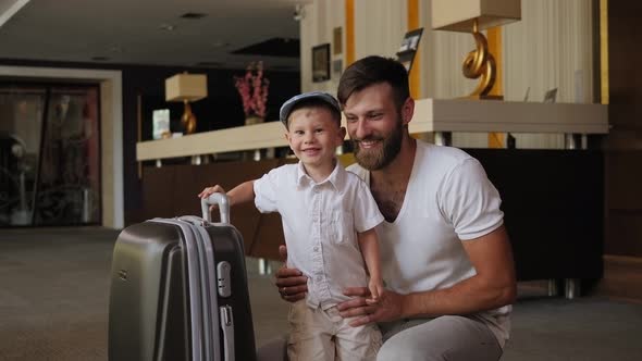 A Happy Bearded Father with a Little Son in a Hotel with a Suitcase alt