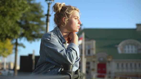 Sideview of Relaxed Attractive Blonde Woman in Denim Jacket Leaning on Fence Blinking As Looking at alt