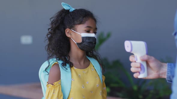 Diverse male teacher measuring temperature of schoolchildren, all wearing face masks alt
