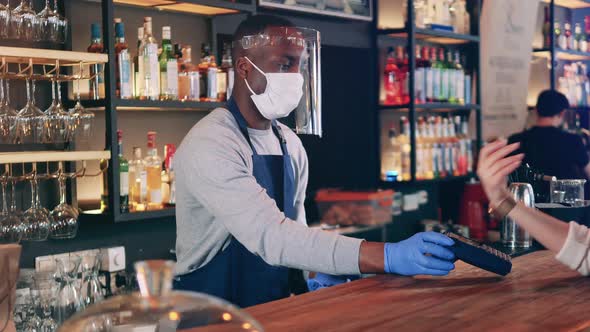 African Ethnicity Waiter in a Face Mask Accepts NFC Payment in the Bar alt
