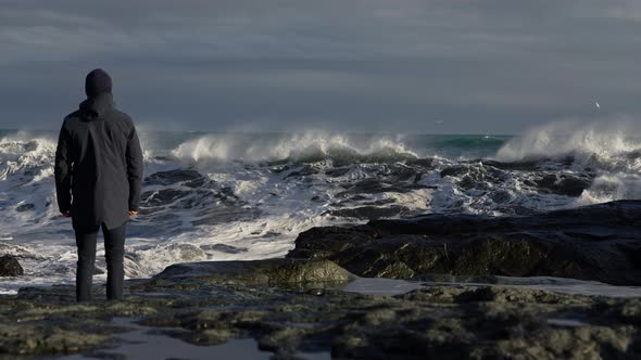 Man Standing On Rocky Shore And Moving To Dodge Crashing Waves alt