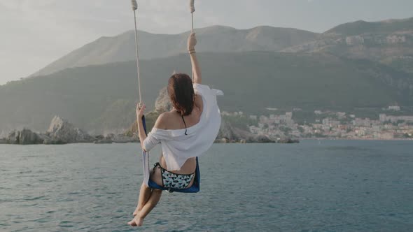 Back view brunette woman in a swimsuit sits on a swing above the sea against mountains alt