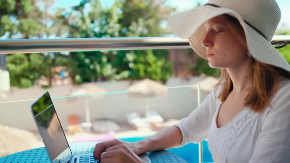 Woman in Hat Works Online on Laptop on Vacation at Hotel with Pool in Summer