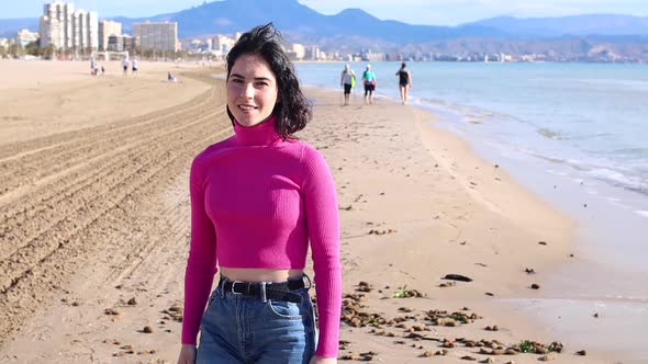 Dressed Young Woman Goes Along the Coastline and Smiling alt