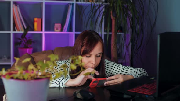 Young Woman Using Smartphone Sitting in Armchair Before Computer in Office alt