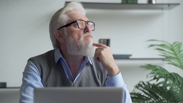 Thoughtfulan Elderly Business Man with a Gray Beard Thinking of Problem Solution Working on Laptop. alt