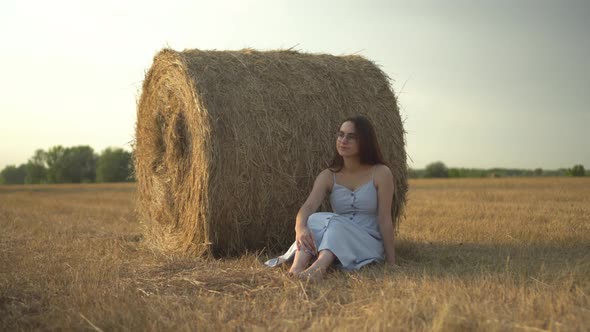 A Young Woman is Sitting Near a Haystack in a Field and a Bee Has Flown to Her alt