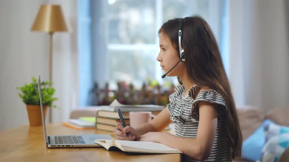 Positive Kid in Headset Writing on Notebook and Using Laptop at Home Studying Online alt