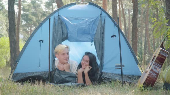 Happy Young Caucasian Couple Lying in Tent in Forest and Smiling at Camera. Portrait of Relaxed alt