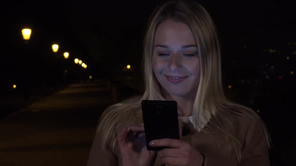 A Young Woman Works on a Smartphone in an Urban Area at Night, Looks Up and Smiles at the Camera alt