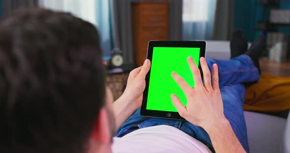 Young Man at Home Resting on a Couch Using with Green Mockup Screen Tablet Computer alt