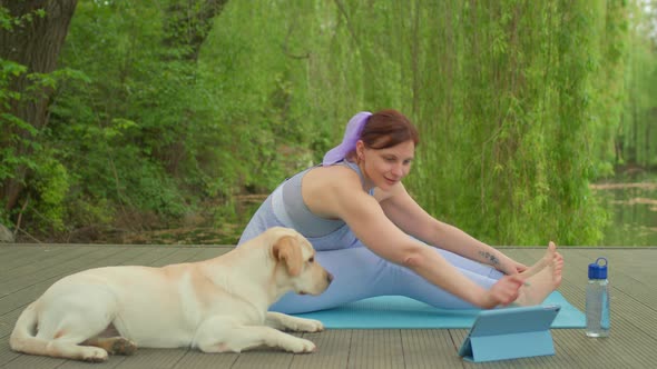 30s Woman Doing Stretching Exercise with Pet Dog Labrador Retriever Lying Near alt