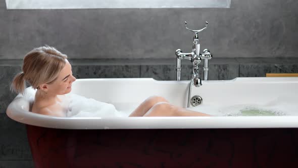Seductive Naked Young Happy Female Taking Bath Surrounded By Foam in Luxury Bathroom Side View alt