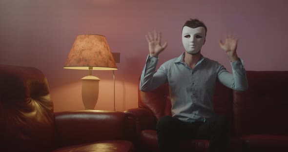 Anonymous man with mask greets with his hand while sitting on the red sofa at home alt