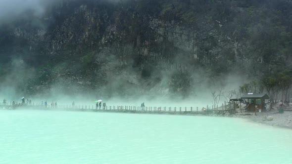 people walking on a bridge with steam rising from sulfur lake and mountain behind, aerial alt