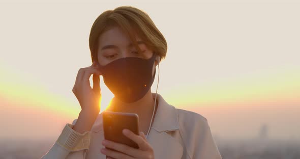 Young Asian woman wearing face mask listening to music from mobile phone outdoors in twilight