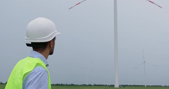 Engineer with beard puts on white helmet, looks at wind turbine generating electricity.