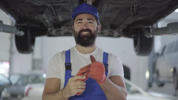Adult Mechanic Takes Out Gloves Standing Under the Lifted Car and Smiling. Bearded Man in Uniform alt