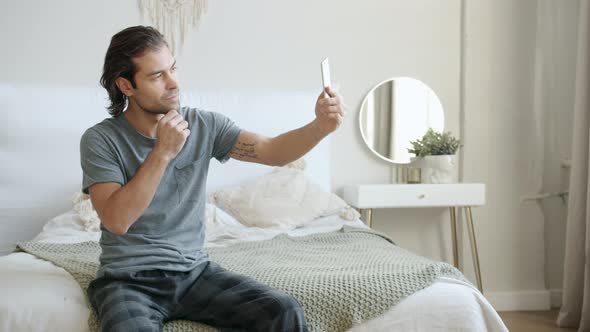 Good-looking Young Man Taking Selfie Using Smartphone Camera Sitting in Bed at Home alt