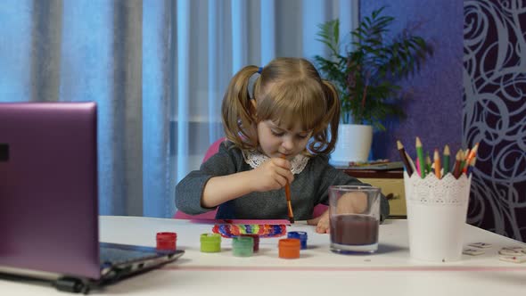 Child Girl Pupil Enthusiastically Drawing with Brushes and Paints Using Laptop Computer at Home alt