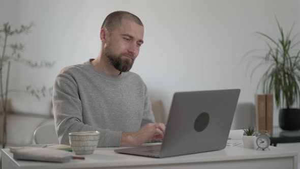 Sleepy male freelancer with beard in a gray jacket works at a laptop at home. alt