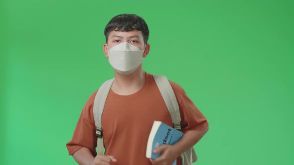 The Front View Of Asian Boy Student Wearing A Mask, Holding A Book And Running On Green Screen alt