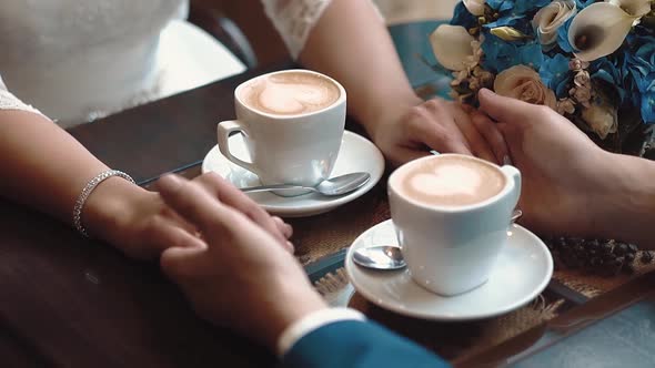 Newlyweds Hold Hands While Sitting in a Cafe, Sitting at a Table on Which There Are Two Mugs of alt