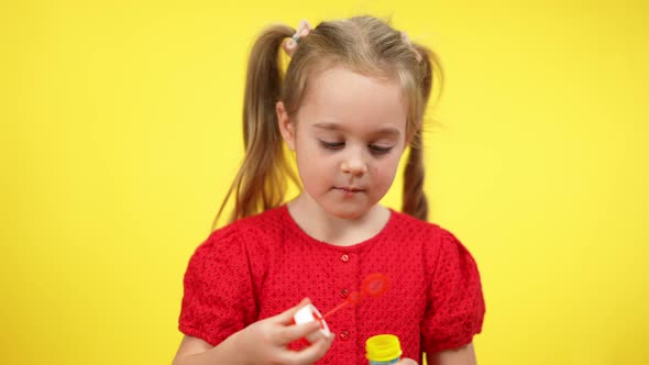 Portrait of Blond Cute Girl with Pigtails Blowing Soap Bubbles at Yellow Background alt
