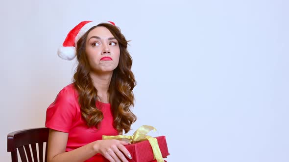 A Beautiful Woman Sits on a Chair in a Christmas Hat with a Gift Shows Disappointed Emotions on a