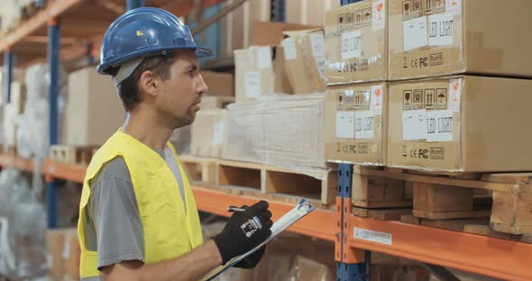 Logistics worker wearing a helmet working in a large warehouse checking inventory alt