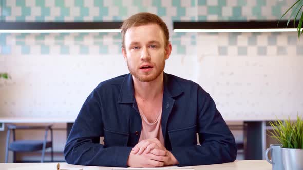 Young Casual Irritated Man Sitting at the Table in Office and Talking to you Aggresively alt